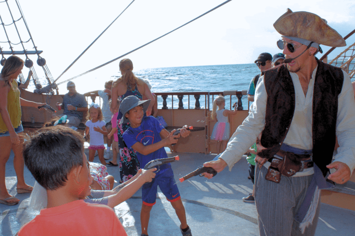 Children playing with toy swords with a pirate actor on a ship deck.