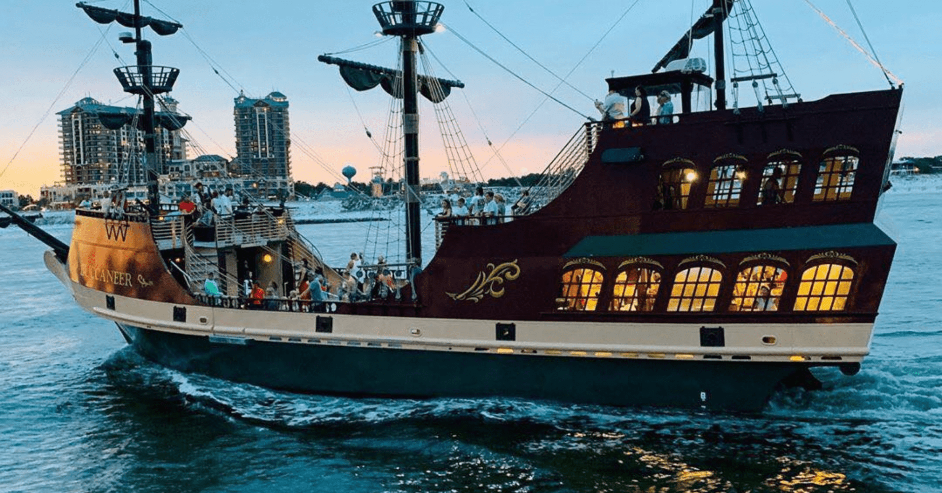 A pirate-themed ship with people onboard sails near city buildings at sunset.