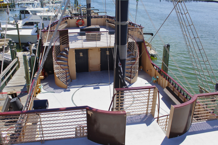 Deck of a docked ship with spiral stairs, mast, and nearby boats in a marina.