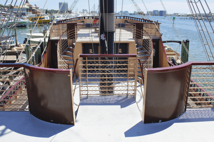 Ship deck with brown railings and steps leading to a higher platform, water and buildings in background.