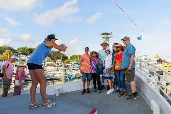 Tourists posing for a group photo on a boat deck, with a woman taking the picture.