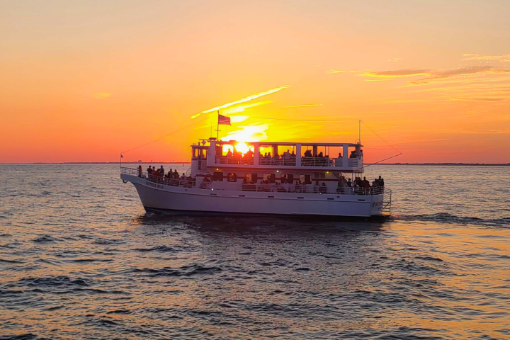 Large boat on ocean at sunset with passengers silhouetted against the orange sky.