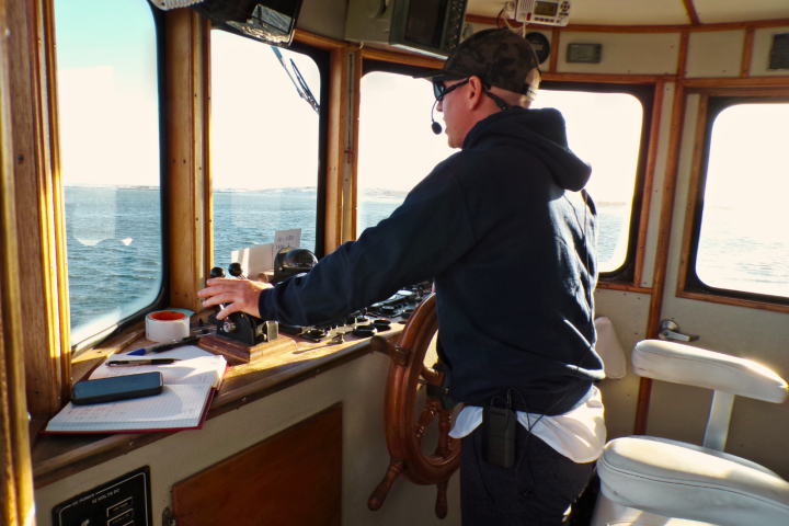 Person steering a boat with a wheel, wearing headset, facing the sea.