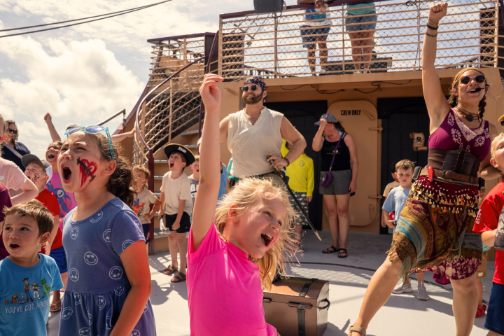Children and adults on a ship deck raise their arms joyfully.
