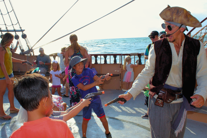 Children and pirate actor playing with toy guns on a ship deck at sea.