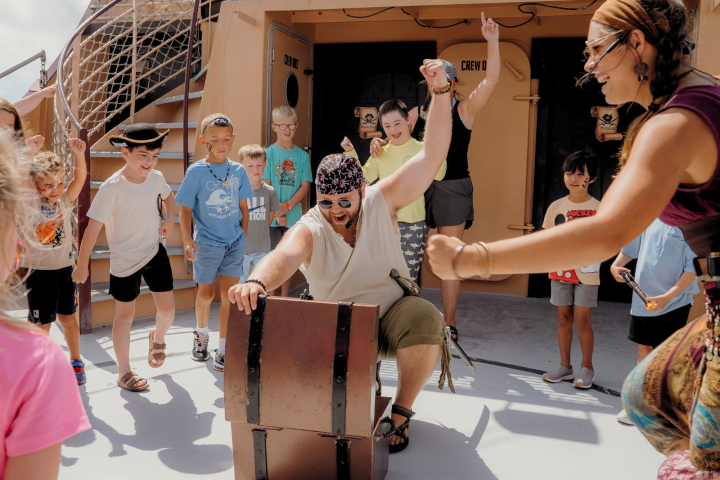 Adults and kids in pirate costumes celebrate around a chest on a ship deck.