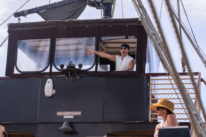 Person pointing from ship's cabin, other person in hat looks on deck.
