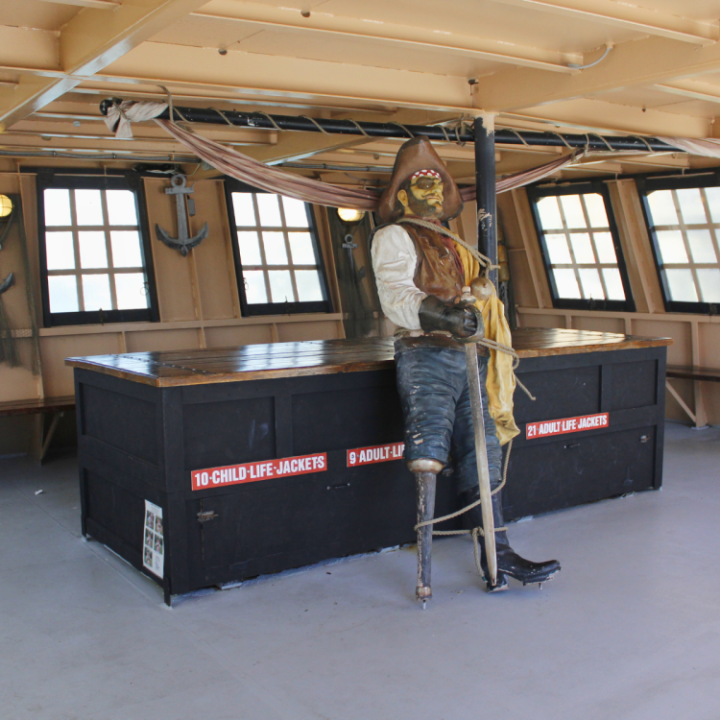 Pirate statue with a peg leg and pirate hat on a ship, near life jacket boxes.