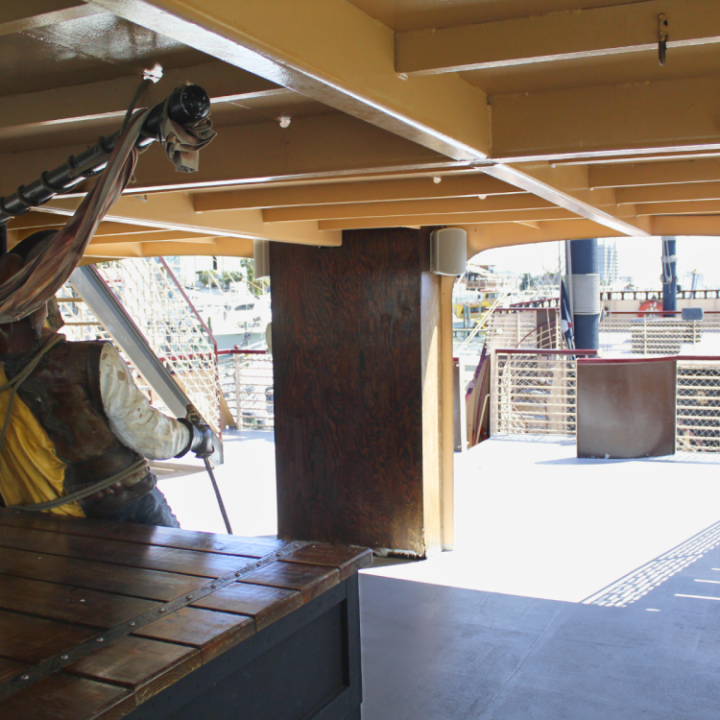 Interior of a vintage ship with figure in historical clothing holding a pole, sunlight filtering through.