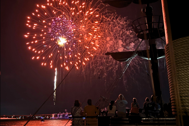 Fireworks bursting in red and purple near a silhouetted ship at night over water.