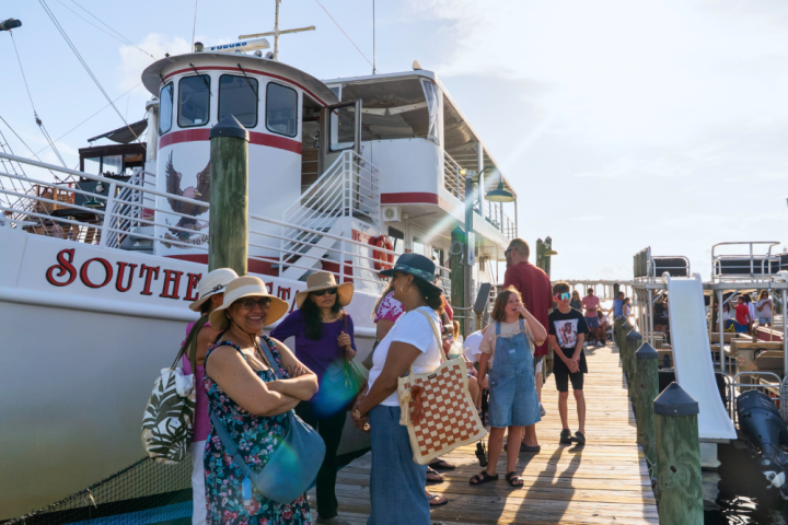 Group of people stand on a dock near a large boat under a sunny sky.