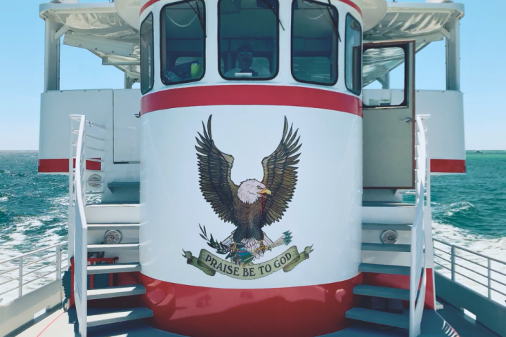 Eagle emblem with 'Praise Be to God' on ship's white and red structure, ocean in background.