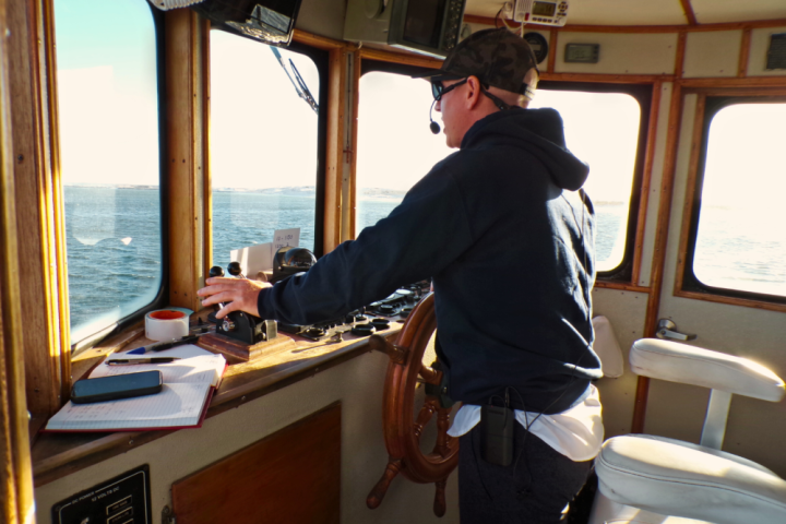 Person steering a boat in a cabin with steering wheel and windows.