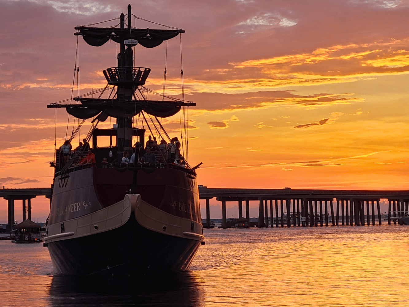 Pirate-style ship at sunset with a bridge in the background over calm water.