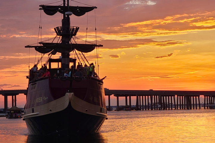 Pirate-style ship at sunset with a bridge in the background over calm water.