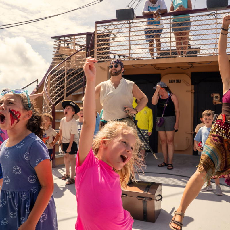 Kids and adults cheer excitedly on a ship's deck under sunny skies.