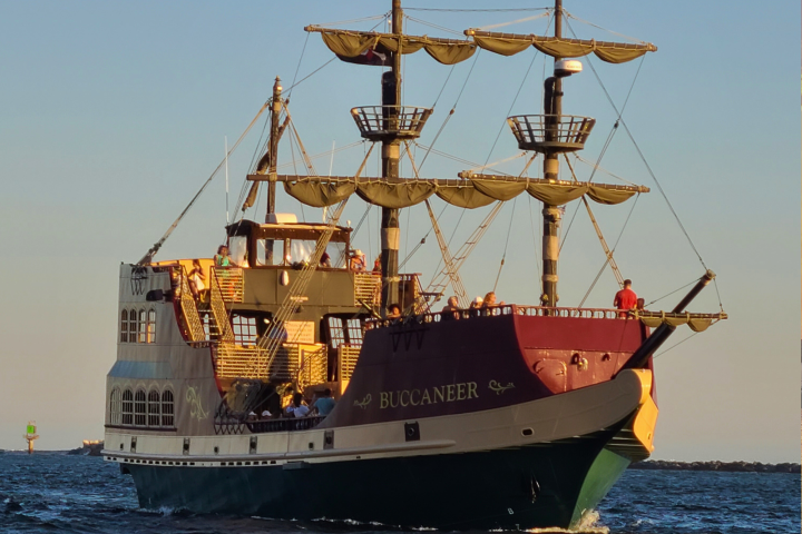 A large pirate ship named Buccaneer sailing on the ocean at sunset.