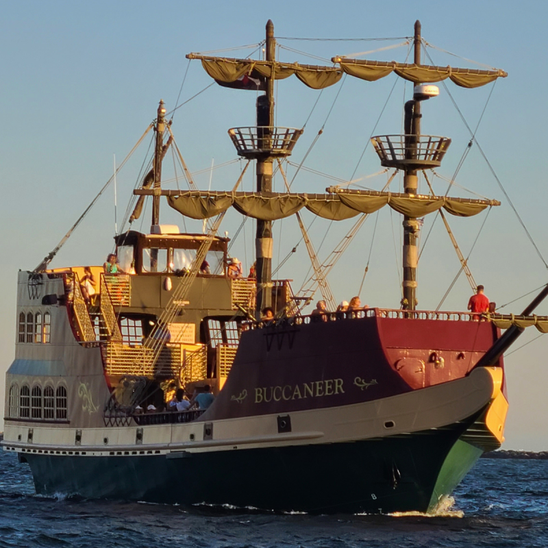 A large pirate ship named Buccaneer sailing on the ocean at sunset.
