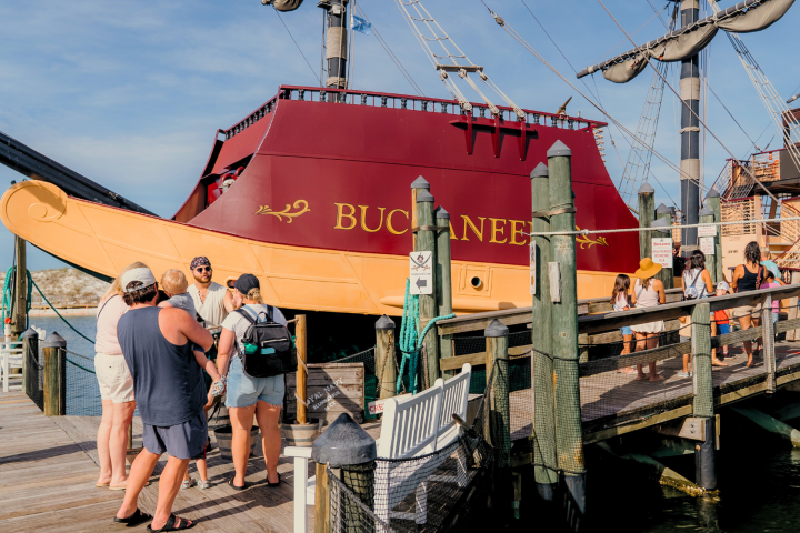 People gather near a docked pirate ship with 'Buccaneer' painted on its side.