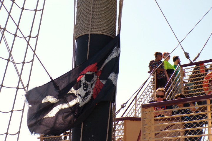Pirate flag on ship mast near people on deck with netting and ropes.