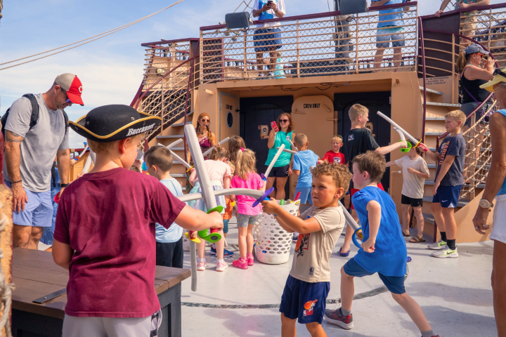 Children play with toy swords on a ship deck, supervised by adults.