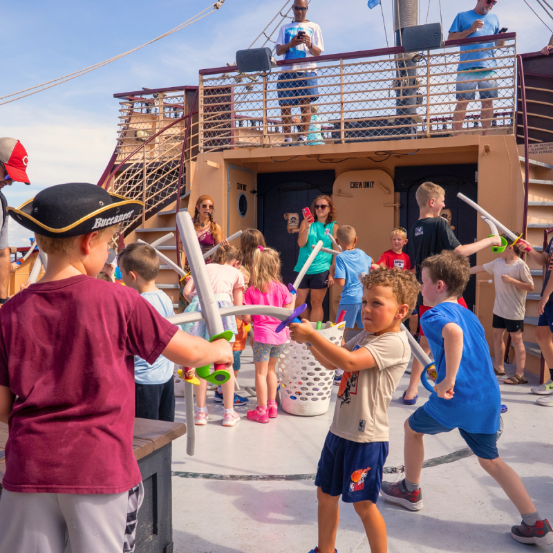 Children play with toy swords on a ship deck, supervised by adults.