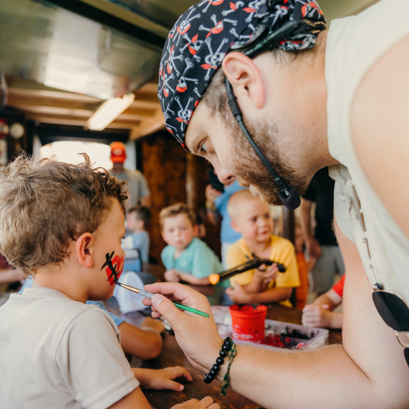 Man painting a child's face in a busy craft area with others around.