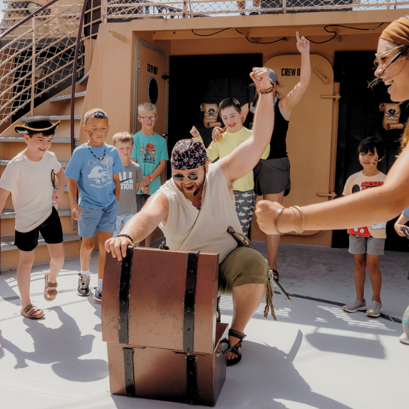 Children cheer as a man dressed as a pirate opens a treasure chest on a ship deck.
